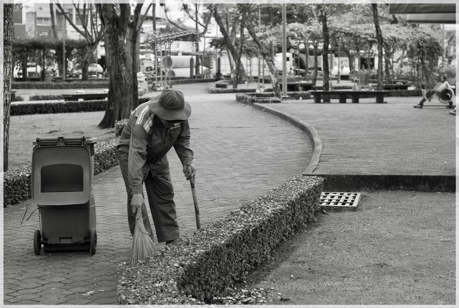 A street scene in daylight showing a worker wearing a traditional conical hat and dark uniform pushing a green waste bin on a paved sidewalk. The worker holds a bamboo broom and is positioned near a l