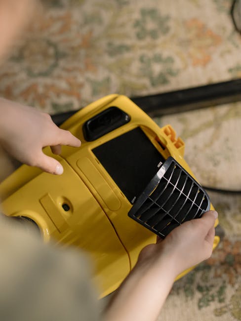 A yellow canister vacuum cleaner with a black flexible hose and metal extension wand is positioned on a decorative area rug in a living room, ready for surface cleaning or deep cleaning. The room feat